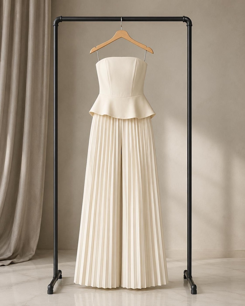 May include: A cream-colored strapless jumpsuit featuring a fitted bodice with a peplum waist and wide-leg trousers with vertical pleats throughout. The garment is displayed on a wooden hanger suspended from a black metal clothing rack.