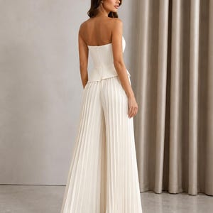 May include: A cream-colored strapless jumpsuit featuring a fitted bodice with a peplum waist and wide-leg trousers with vertical pleats. The garment has a clean, structured silhouette and a smooth finish, showcasing a cohesive monochromatic design.