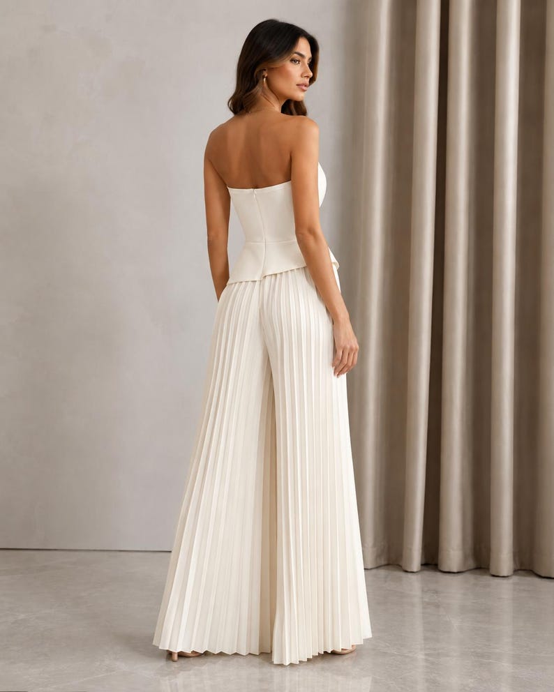 May include: A cream-colored strapless jumpsuit featuring a fitted bodice with a peplum waist and wide-leg trousers with vertical pleats. The garment has a clean, structured silhouette and a smooth finish, showcasing a cohesive monochromatic design.