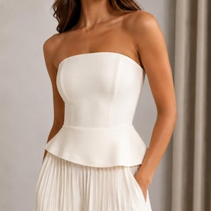 May include: A strapless white peplum top featuring a structured bodice with vertical seams, paired with matching white pleated trousers that include side pockets. The ensemble is crafted from a smooth, solid-colored fabric with a clean finish.