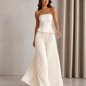 May include: A cream-colored strapless jumpsuit featuring a fitted bodice with a peplum waist and wide-leg trousers with vertical pleats. The garment is constructed from a smooth, solid fabric and extends to the floor, creating a long, flowing silhouette.