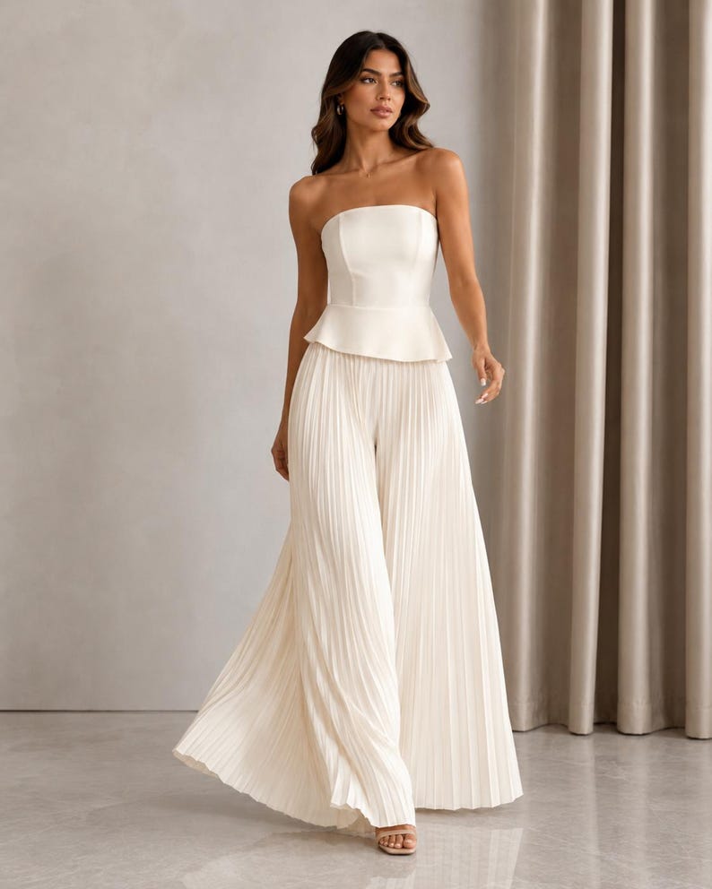 May include: A cream-colored strapless jumpsuit featuring a fitted bodice with a peplum waist and wide-leg trousers with vertical pleats. The garment is constructed from a smooth, solid fabric and extends to the floor, creating a long, flowing silhouette.