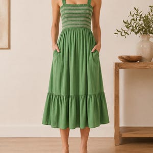 May include: A midi-length green dress featuring a square neckline, wide shoulder straps, and a smocked bodice with white horizontal stitching. The skirt has a gathered waist, side pockets, and a single tiered ruffle at the hemline.