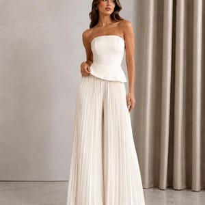 May include: A strapless white peplum top paired with matching floor-length wide-leg trousers. The trousers feature a vertical pleated texture throughout, creating a flowing silhouette. The ensemble is a solid off-white color with a clean, structured bodice.