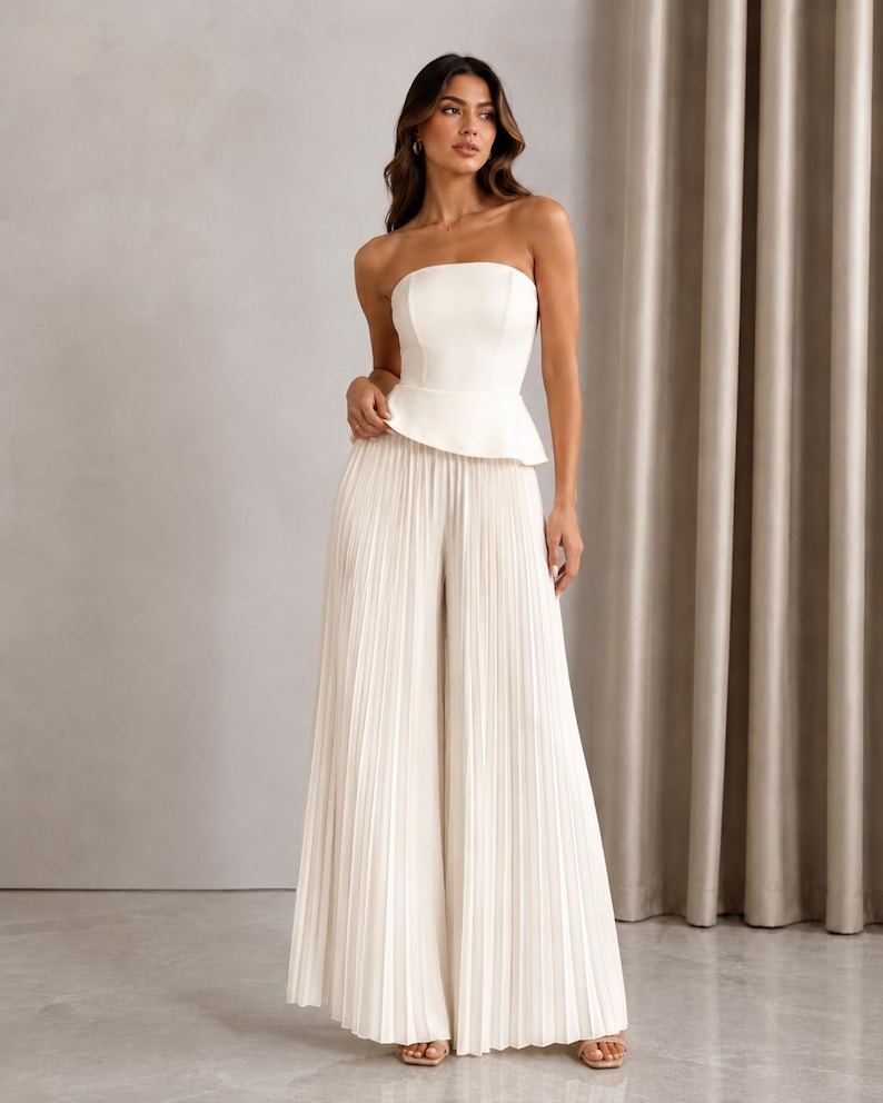 May include: A strapless white peplum top paired with matching floor-length wide-leg trousers. The trousers feature a vertical pleated texture throughout, creating a flowing silhouette. The ensemble is a solid off-white color with a clean, structured bodice.