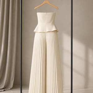 May include: A cream-colored strapless jumpsuit featuring a fitted bodice with a peplum waist and wide-leg trousers with vertical pleats throughout. The garment is suspended on a wooden hanger from a black metal clothing rack.