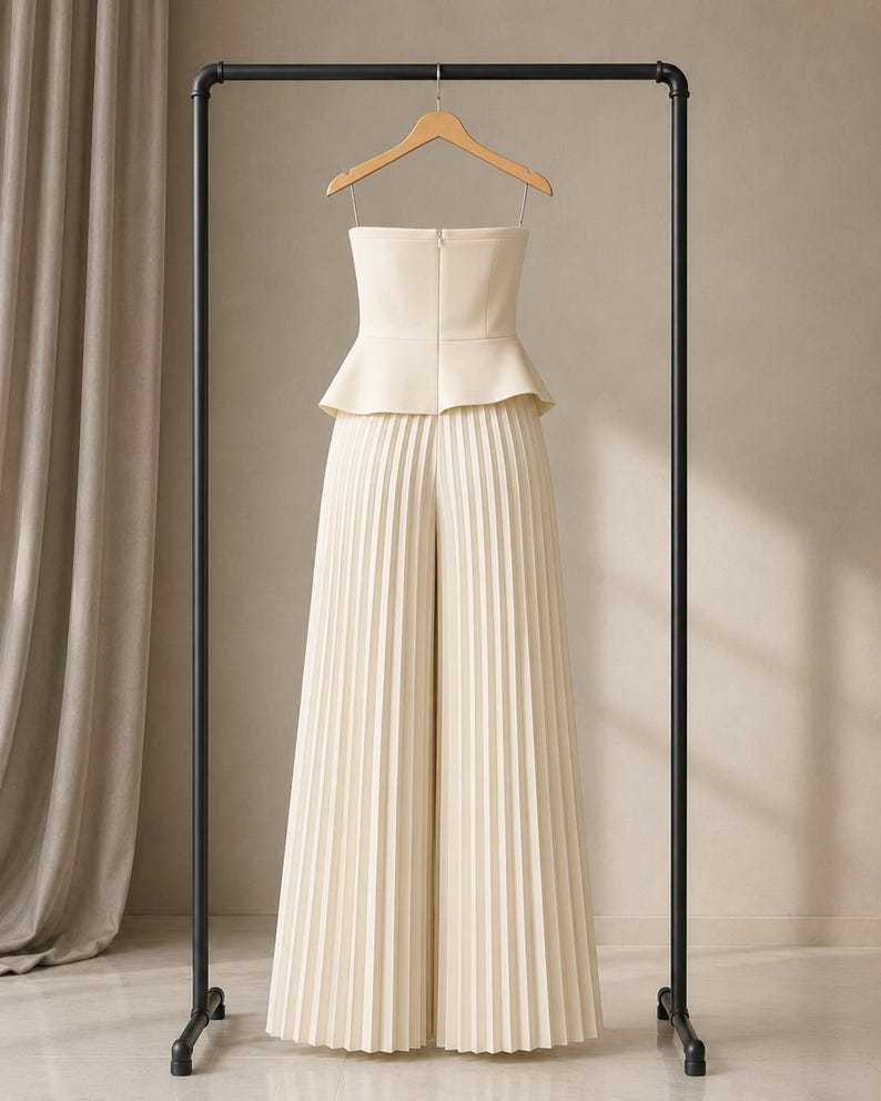 May include: A cream-colored strapless jumpsuit featuring a fitted bodice with a peplum waist and wide-leg trousers with vertical pleats throughout. The garment is suspended on a wooden hanger from a black metal clothing rack.