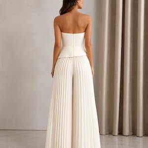 May include: A white strapless jumpsuit featuring a fitted bodice with a peplum waist detail and wide-leg trousers with vertical pleats throughout. The garment has a clean, solid white color and a structured silhouette with a back zipper closure.