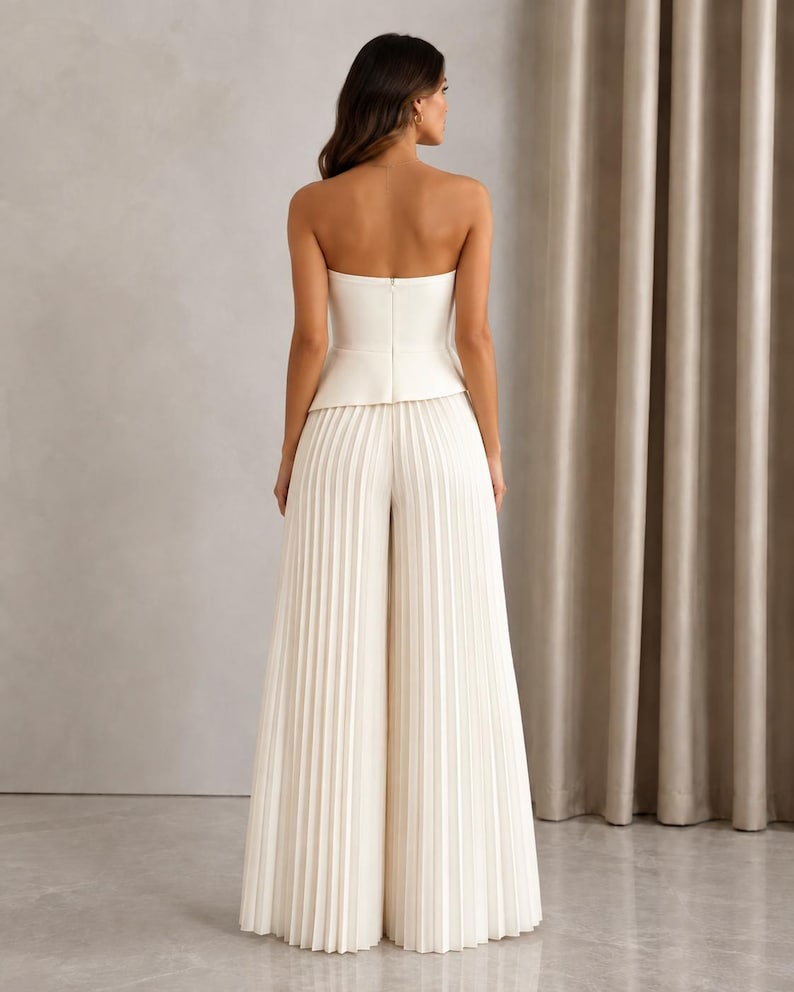 May include: A white strapless jumpsuit featuring a fitted bodice with a peplum waist detail and wide-leg trousers with vertical pleats throughout. The garment has a clean, solid white color and a structured silhouette with a back zipper closure.