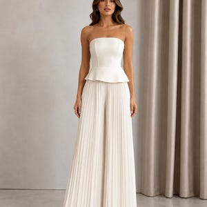 May include: A white strapless jumpsuit featuring a fitted bodice with a peplum waist detail and wide-leg trousers with vertical pleats throughout the length of the pants. The garment is a solid off-white color with a clean, structured silhouette.