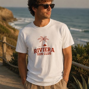 Könnte beinhalten: Ein weißes kurzärmeliges T-Shirt mit einem Grafikdruck in rostorangefarbener Tinte. Das Design zeigt eine Palme über einer untergehenden Sonne, wobei der Text RIVIERA SUN CLUB in einer fetten, großgeschriebenen Serifenschrift auf der Brust steht.