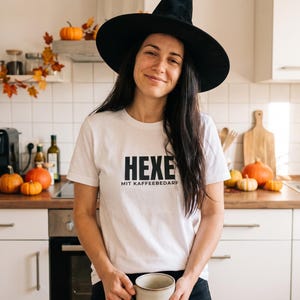 May include: A white short-sleeved t-shirt featuring the black text "HEXE MIT KAFFEEBEDARF" printed on the chest. The shirt is paired with a black pointed witch hat and a plain, light-coloured ceramic mug held in both hands.