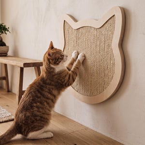 May include: A wall-mounted cat scratching post shaped like a cat head, featuring a light-coloured wooden frame and a central panel covered in textured tan sisal rope. A ginger and white tabby cat is standing on its hind legs, scratching the surface.