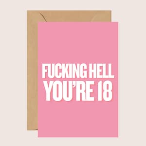 May include: A rectangular greeting card featuring a solid pink background with bold white text stating FUCKING HELL YOU'RE 18. The card is placed in front of a plain brown envelope, highlighting the contrast between the vibrant pink and the neutral paper.