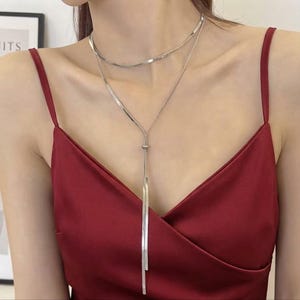 Op de afbeelding: Een zilverkleurige gelaagde ketting bestaande uit een platte slangenketting als choker en een bijpassende hangende ketting in lariat-stijl. De ketting wordt gedragen over een donkerrode satijnen top met dunne spaghettibandjes en een V-hals.