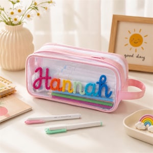 Peut inclure: Une trousse à crayons rectangulaire et transparente avec une bordure rose et une poignée latérale. Le devant présente le prénom Hannah en lettres cursives colorées et duveteuses. L'intérieur contient des stylos et des crayons visibles.
