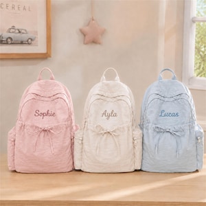 May include: Three backpacks in light pink, cream, and light blue, each featuring a front pocket with a drawstring tie and a name embroidered in cursive script: Sophie on the pink, Ayla on the cream, and Lucas on the light blue backpack.