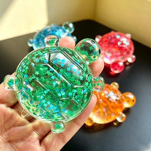 May include: A clear green turtle-shaped figurine filled with small blue star-shaped glitter. The turtle has a smooth, rounded shell and four small flippers. Other similar turtle figurines in blue, red, and orange are visible in the background.