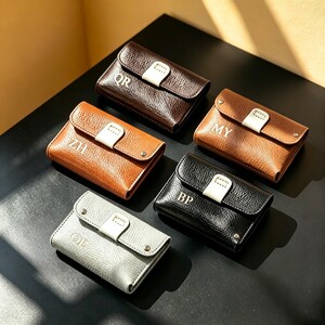 May include: Five small rectangular wallets in dark brown, light brown, black, and light gray leather with a textured finish. Each wallet features a white tab closure, silver-tone metal snap buttons, and two-letter monograms: QR, ZH, MY, BP, and OJE.