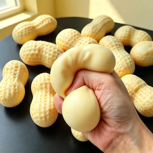 Può includere: Una collezione di giocattoli antistress a forma di arachide, di colore beige chiaro, con superfici testurizzate simili a gusci. Uno dei giocattoli viene compresso, mostrando il suo materiale flessibile e la forma arrotondata, mentre gli altri mantengono la loro forma originale.