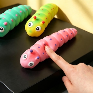 May include: Three segmented caterpillar-shaped sensory toys in pink, yellow-green, and bright green. Each toy features two antennae, large white eyes with black pupils, and small purple or pink polka dots along its flexible, rounded body.