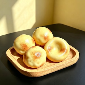 May include: Four smooth, round, pale yellow objects with brown speckled markings arranged on a square wooden tray with rounded corners. The objects have a slightly irregular, organic shape and a glossy finish, resting together on the light wood surface.