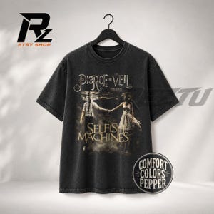 May include: A charcoal grey short-sleeve t-shirt featuring a graphic print of a person in a dress and a person in a button-down shirt standing in water. The text reads Pierce the Veil Presents Selfish Machines. A circular badge says Comfort Colors Pepper.