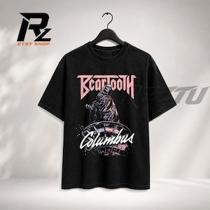 Puede incluir: Una camiseta negra de manga corta con un estampado gráfico que incluye el texto Beartooth en rosa y Columbus en blanco. La ilustración central muestra una figura encapuchada de pie sobre una estructura circular en la tela oscura.