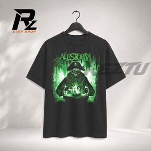 May include: A black short-sleeve t-shirt featuring a graphic print of a skeletal pirate wearing a hat, with the text "ALESTORM" above. The pirate has a hook for a hand and is interacting with a glowing green object in a dark, cavernous setting.