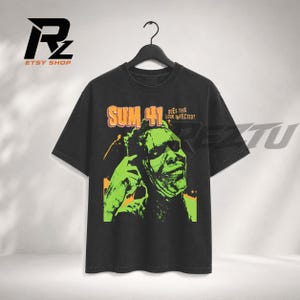 Op de afbeelding: Een zwart t-shirt met korte mouwen en een grafische print van een groen getint gezicht met de tekst "SUM 41" en "DOES THIS LOOK INFECTED?" in oranje en wit. Het shirt hangt aan een zwarte kledinghanger tegen een neutrale achtergrond.