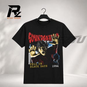 May include: A black short-sleeve t-shirt featuring a graphic print of a person with hands pressed against a surface. The text reads SOUNDGARDEN, SUPERUNKNOWN, FELL ON BLACK DAYS, and 1994. The design uses red, yellow, and blue tones on a black background.