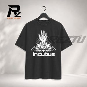 Puede incluir: Una camiseta negra de manga corta con un estampado gráfico blanco de una mano haciendo un gesto sobre una flor de loto con un símbolo Om en el centro. La palabra incubus está impresa en texto blanco debajo del diseño de loto al frente.