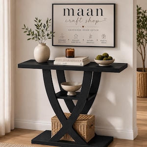 May include: A black console table with an X-shaped base and a lower shelf holding a woven basket. On top sits a beige vase with greenery, a brown candle, two stacked books, and a bowl of green spheres. A sign reads "maan craft shop".