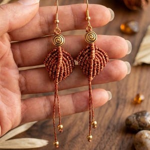 May include: A pair of dangling earrings featuring a brown macrame leaf-shaped design suspended from gold-toned metal hooks. Each earring includes a circular gold-toned accent with a spiral pattern and two thin cords ending in small gold-toned beads.
