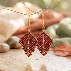 Puede incluir: Un par de pendientes colgantes con cordón de macramé color óxido tejido en forma de diamante. Cada pendiente está adornado con una pequeña cuenta de tono dorado en la parte superior e inferior, suspendido de ganchos de metal dorado.