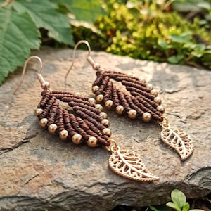 Könnte beinhalten: Ein Paar braune Makramee-Ohrringe mit blattförmigem Design und kleinen goldfarbenen Perlen an den Rändern. Jeder Ohrring ist mit einem baumelnden, blattförmigen Anhänger aus goldfarbenem Metall und einem einfachen Ohrhaken versehen.