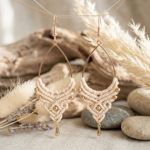 Peut inclure: Une paire de boucles d'oreilles en forme de goutte composées de corde macramé couleur crème tissée en motif géométrique. Chaque boucle est ornée de petites perles en métal doré et d'une breloque goutte dorée suspendue à un crochet doré.
