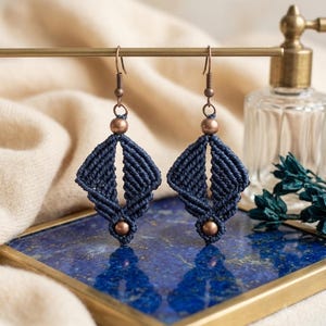 Peut inclure: Une paire de boucles d'oreilles en macramé bleu marine au design géométrique en forme de losange. Chaque boucle est ornée d'une perle métallique cuivrée en haut et en bas, suspendue à un crochet en métal cuivré assorti pour oreilles percées.
