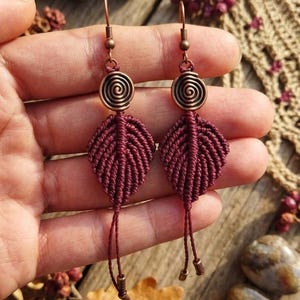 Könnte beinhalten: Ein Paar Ohrhänger mit dunkelroten, blattförmigen Makramee-Anhängern, die an kupferfarbenen Spiral-Metallanhängern und passenden Ohrhaken hängen, mit dünnen Kordelenden an der Basis jedes Blattes.