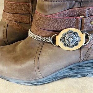 May include: A pair of brown leather wedge boots featuring a textured leather strap across the ankle. The strap is adorned with a silver-toned metal chain and a central circular medallion with a floral pattern set on a light tan leather backing.