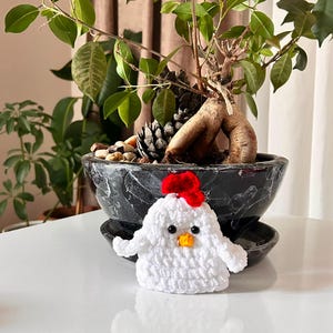 May include: A small, white crocheted chicken figurine featuring a bright red comb on top, a small yellow beak, and two black bead eyes. The handcrafted textile ornament has a rounded body shape with two small wings extending from the sides.