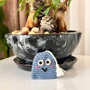 May include: A small, blue, crocheted ghost-shaped ornament with large black and white plastic eyes, a stitched black V-shaped mouth, and pink embroidered cheeks. A white yarn tassel hangs from the top corner of the blue crocheted figure.