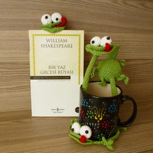 Peut inclure: Un marque-page grenouille en crochet vert avec des yeux blancs et une langue rouge posé sur un livre intitulé William Shakespeare Bir Yaz Gecesi Rüyasi, accompagné d'une tasse noire à motif floral coloré et d'un dessous de verre grenouille.