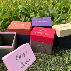 May include: A collection of square, multi-colored plastic containers with lids. The lids feature text including Rooted in Michigan, grower, dirt therapy, Seeds, and Gabby's Garden. Some containers have embossed plant icons on the sides.