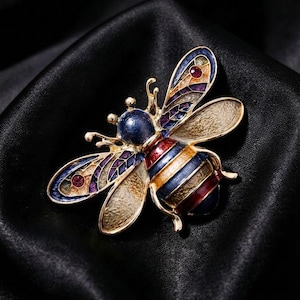 May include: A bee-shaped brooch featuring gold-tone metal, blue, red, and yellow enamel stripes on the body, and wings detailed with purple, blue, and red accents including small embedded crystals. Text in the corner reads PREMIUM ADORNMENTS.