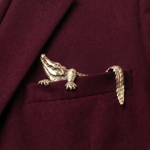 Op de afbeelding: Een goudkleurige metalen krokodillenbroche bevestigd aan de borstzak van een donkerbordeauxrode blazer. De kop en voorpoten van de krokodil komen uit de zak, terwijl de gestructureerde staart over de bovenrand van de zakstof buigt.