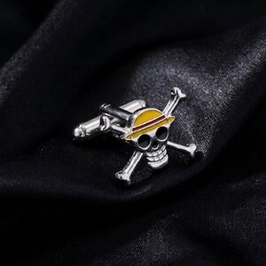 Puede incluir: Un gemelo de metal plateado con un diseño de calavera y huesos cruzados. La calavera lleva un sombrero de paja amarillo con una banda roja. El gemelo tiene un acabado metálico pulido y un mecanismo de cierre de barra en T en la parte posterior.