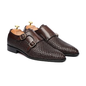Pode incluir: Um par de sapatos de fivela dupla castanho-escuro com uma textura de couro entrançado na parte inferior e couro liso na zona da tira superior. Cada sapato inclui duas fivelas de metal dourado e uma forma de madeira.