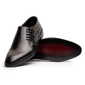May include: A black leather dress shoe featuring a smooth pointed toe with decorative perforations, a side panel with a grid-like embossed texture, and black laces. The shoe is positioned to reveal a dark red sole with a black outer edge.