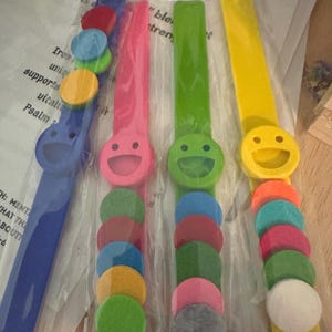 Puede incluir: Cuatro pulseras de silicona de tipo slap en azul, rosa, verde y amarillo. Cada pulsera presenta un icono de cara sonriente circular y varios accesorios pequeños, coloridos y circulares de fieltro en tonos como rojo, naranja, azul y blanco.